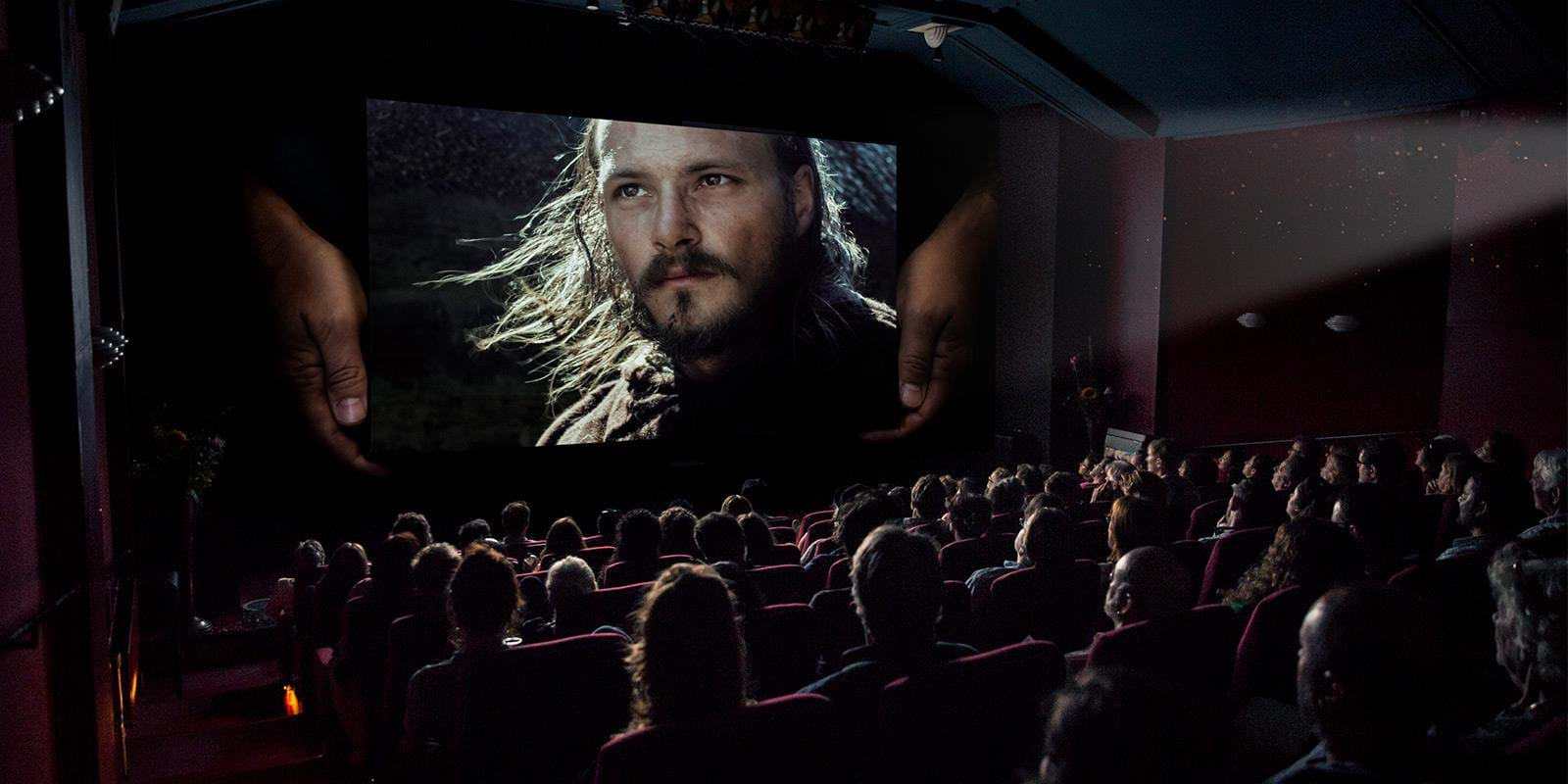 Biografsal fyldt med mennesker hvor kæmpe hænder holder lærred